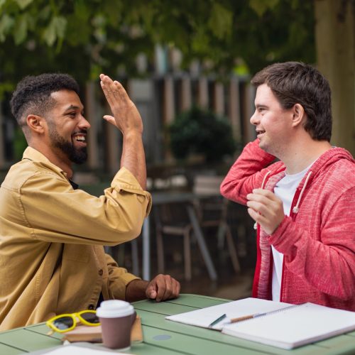 Young man with Down syndrome with his mentoring friend sitting outdoors in cafe high fiving.
